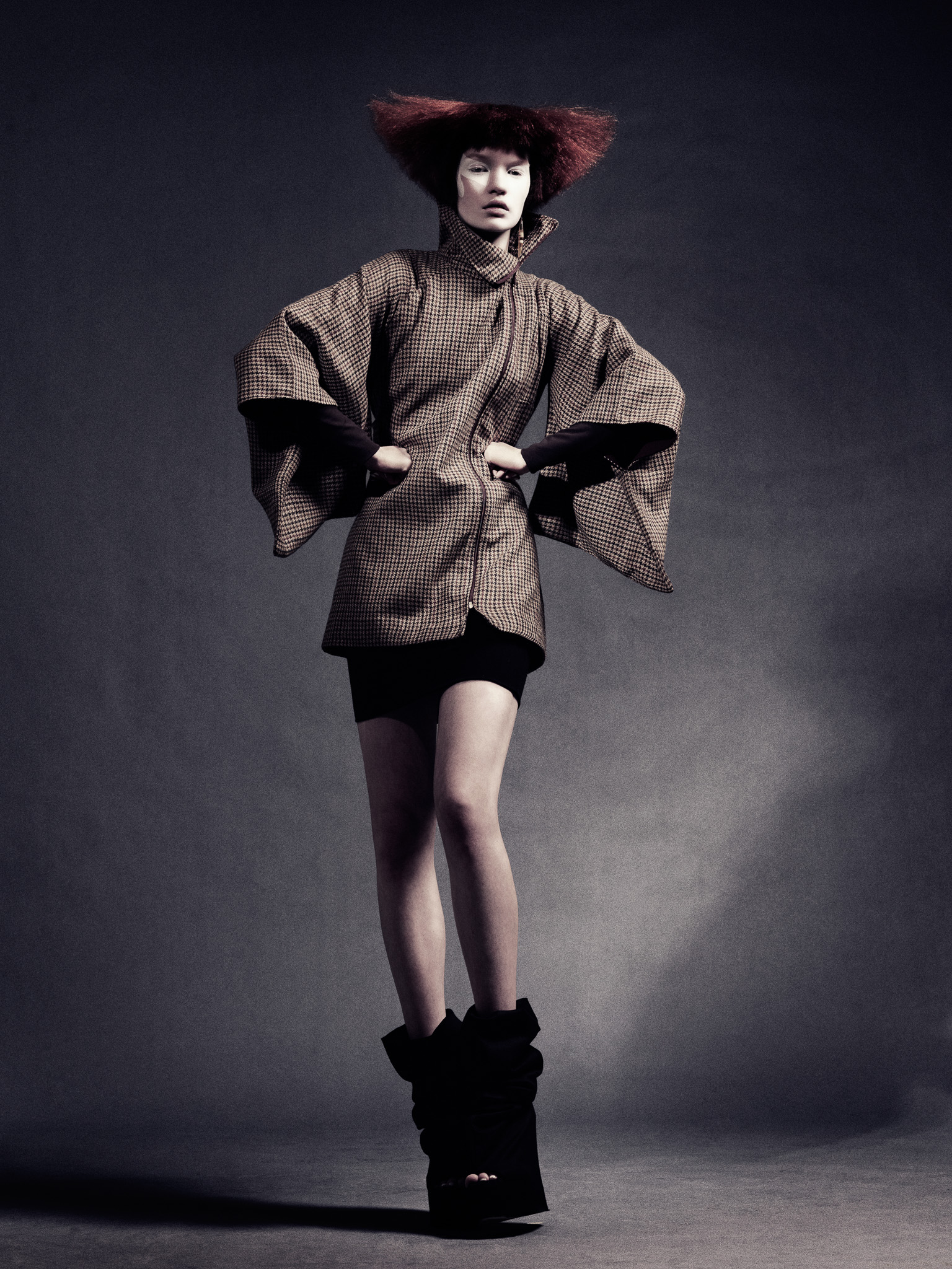 Model wearing a checked structured jacket with wide sculptural sleeves against a dark backdrop
