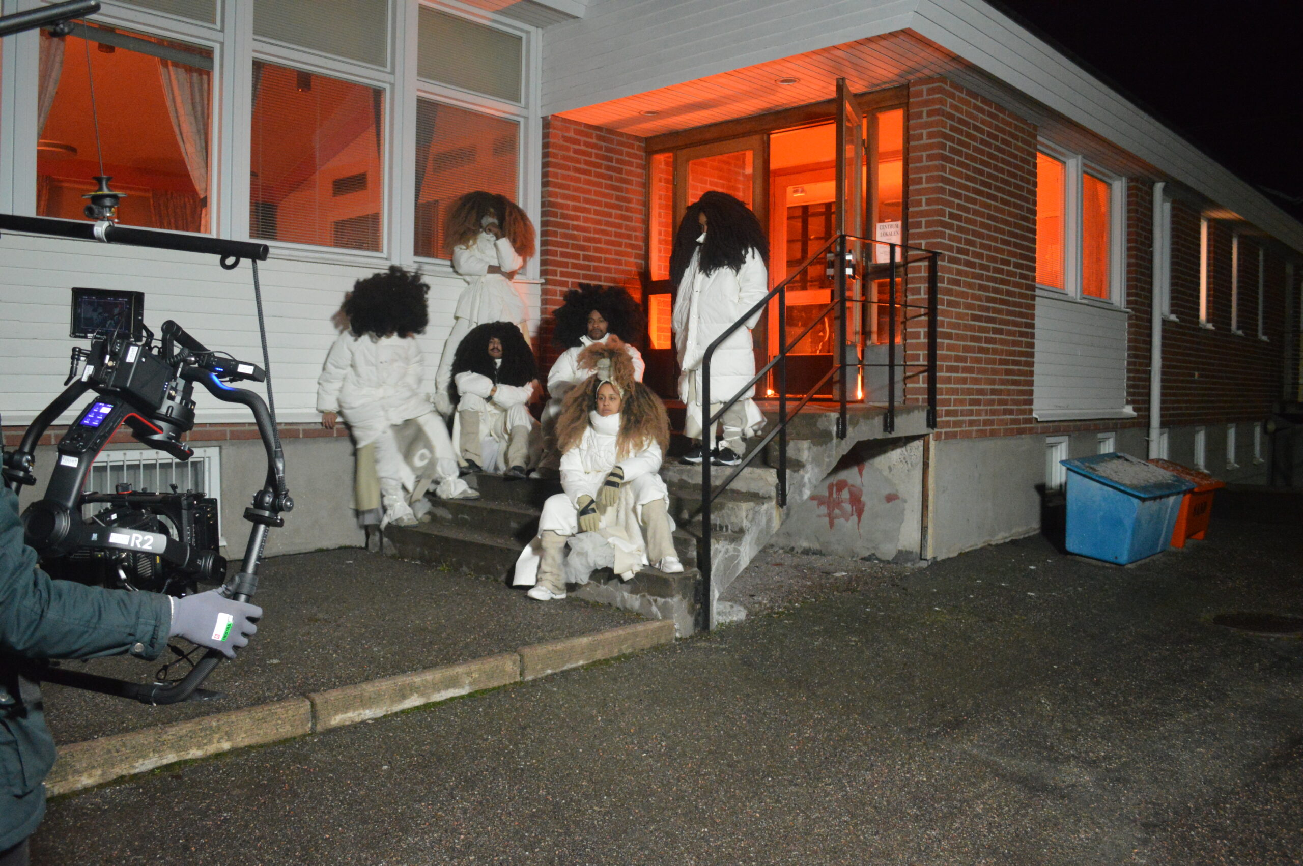 FutureBrownSpace performers in white outfits on the steps of a building during a night shoot, with a camera rig in the foreground.