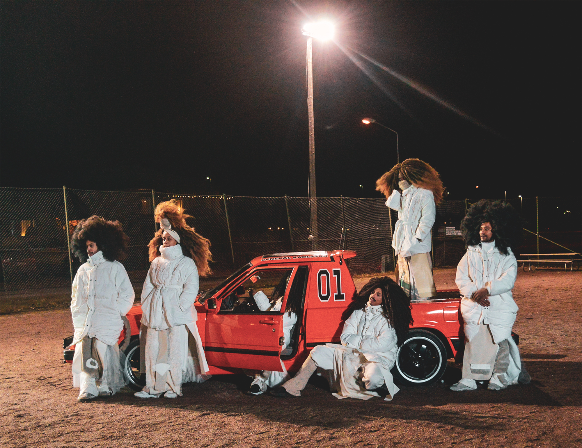 FutureBrownSpace performers in layered white outfits gathered around a red car at night.