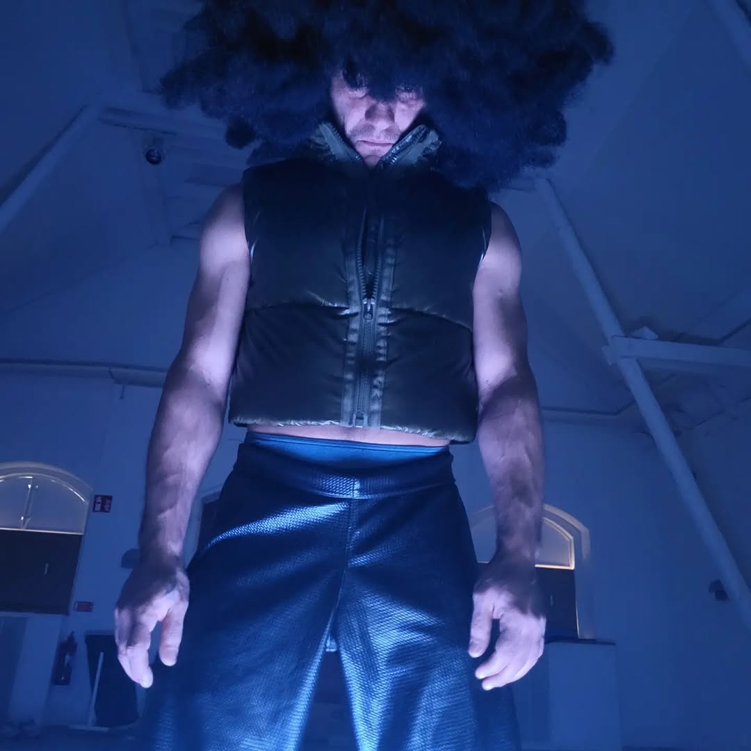 John-Paul Zaccarini in blue light, wearing a large dark afro and sleeveless zippered vest.