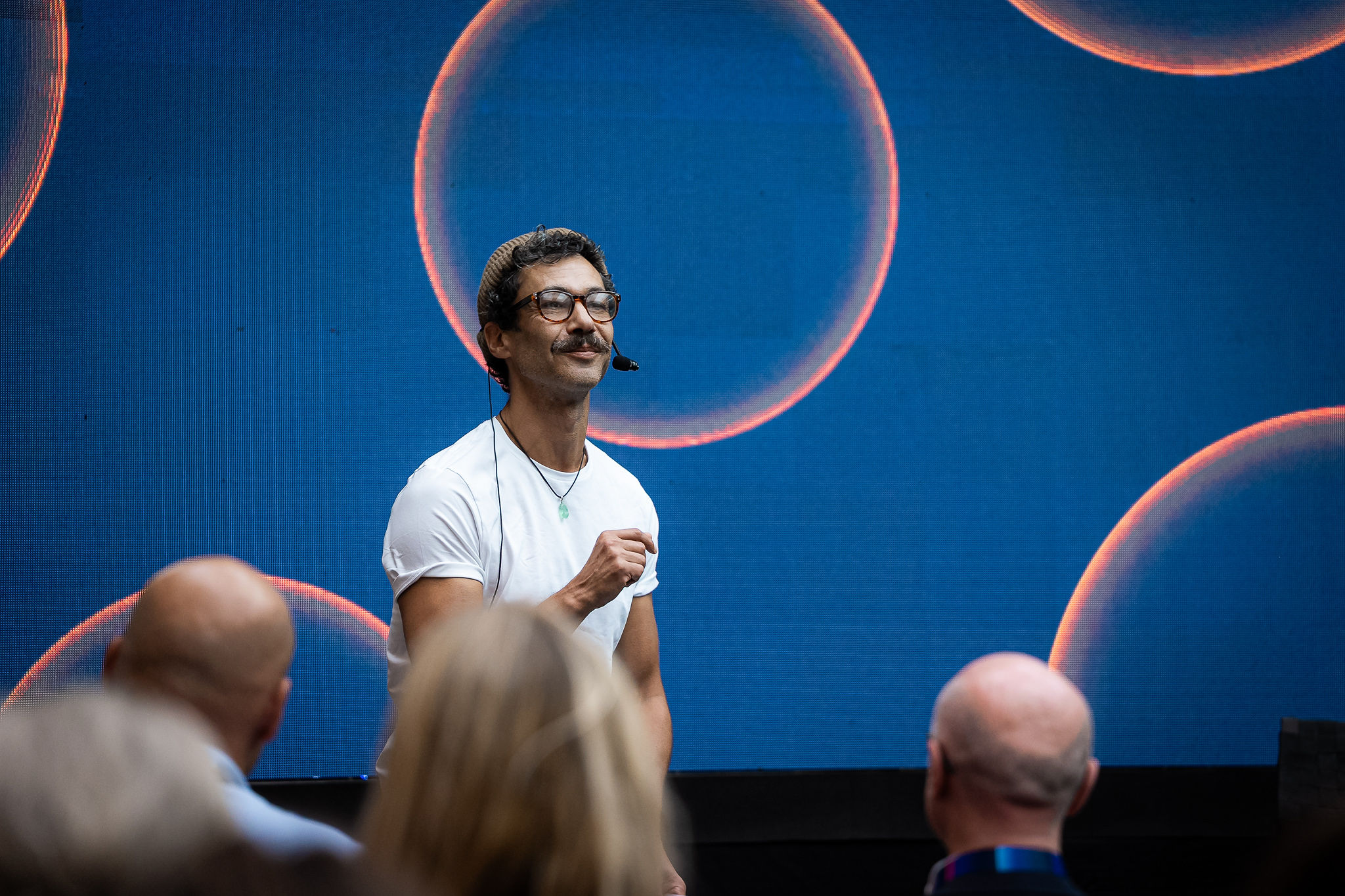 John-Paul Zaccarini speaking onstage in a white T-shirt before a blue screen with circular light graphics.